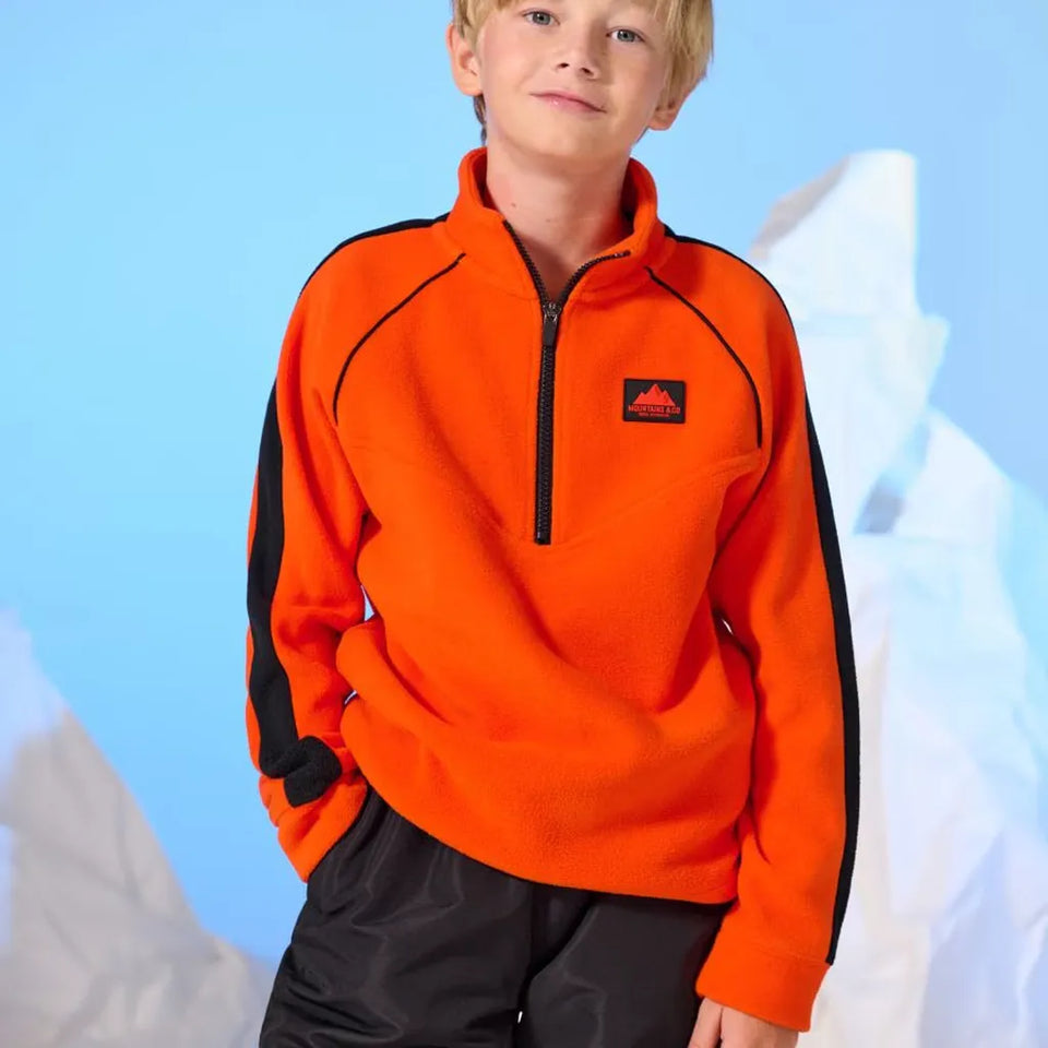 A young boy wearing a bright orange quarter-zip fleece pullover with black side panels and a small red logo patch on the chest, posed against a light blue backdrop with white mountain silhouettes.