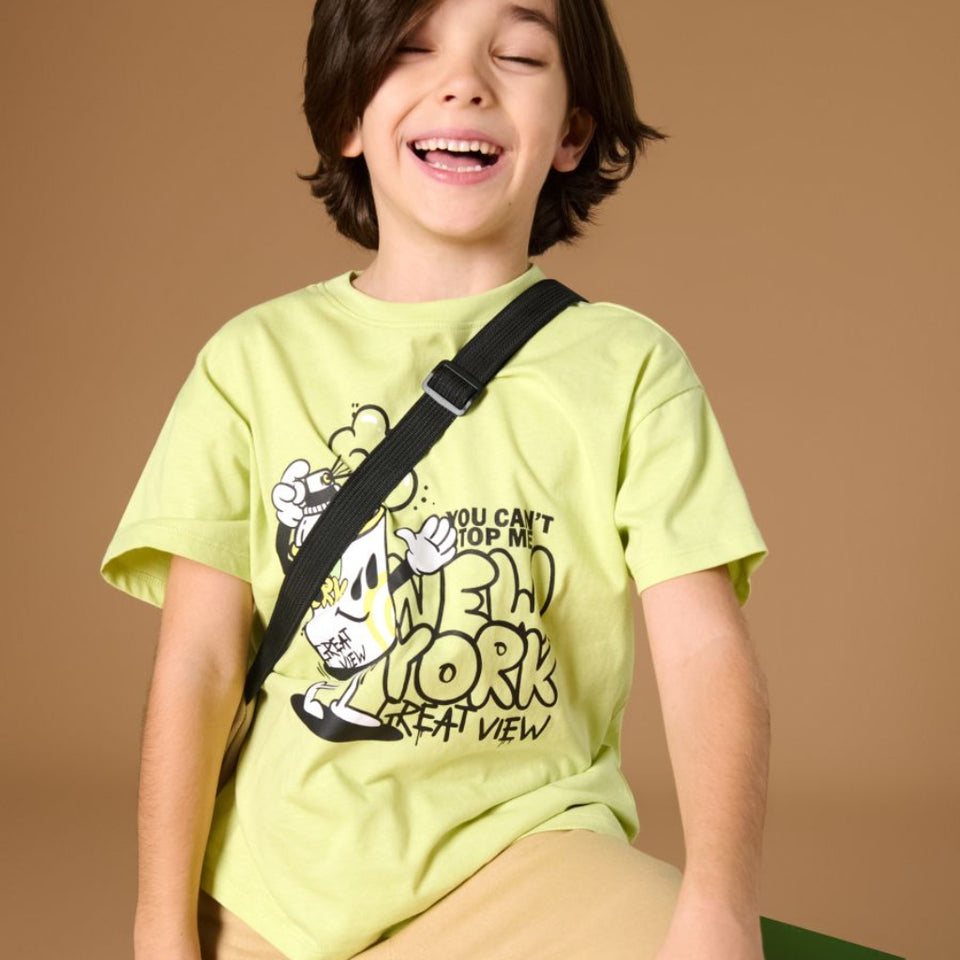 A young boy with dark hair sits smiling on a green stool, wearing a light yellow-green graphic t-shirt with a black strap across his chest and beige pants, set against a solid brown studio background.