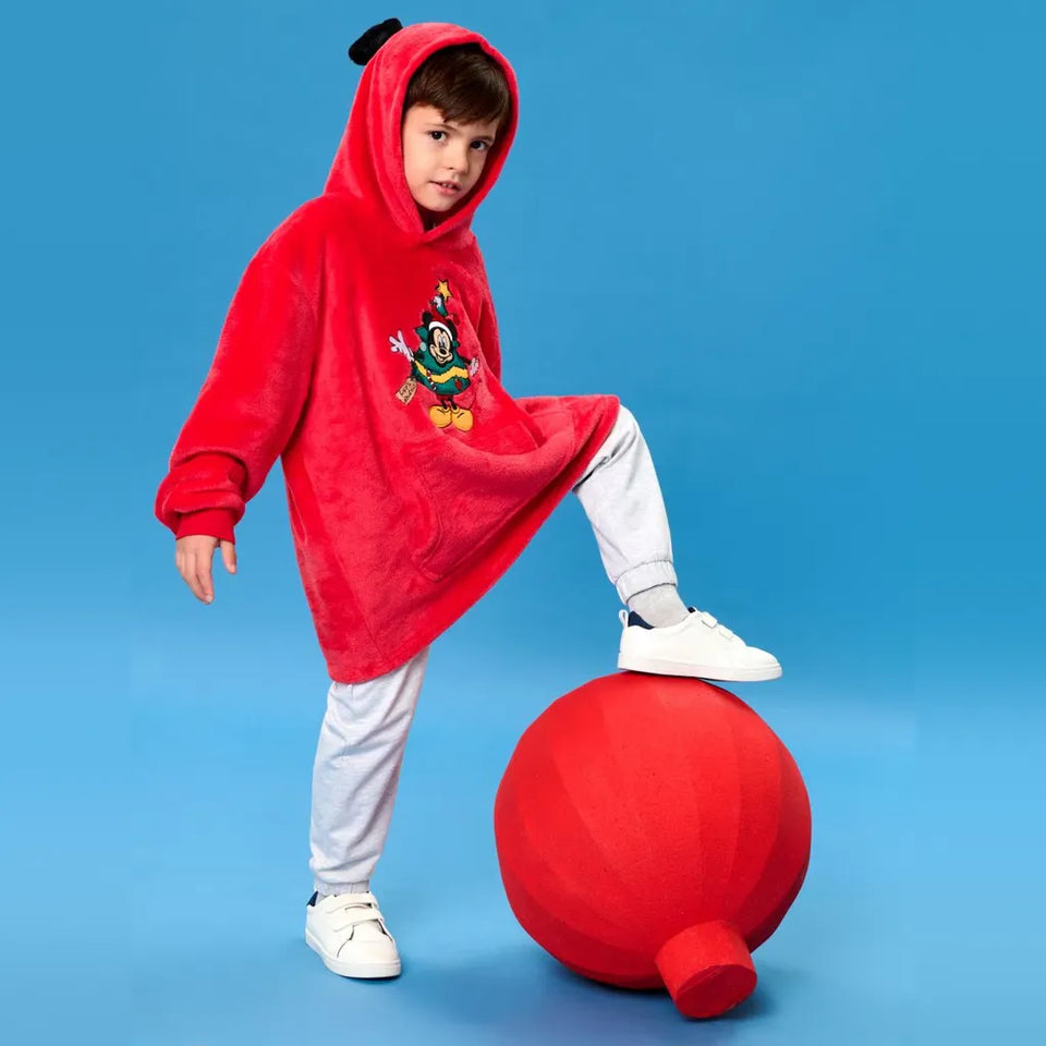 A child wearing a plush red hooded costume with a cartoon character on the front, white pants, and white sneakers, standing on a large red spherical prop against a solid blue studio background.