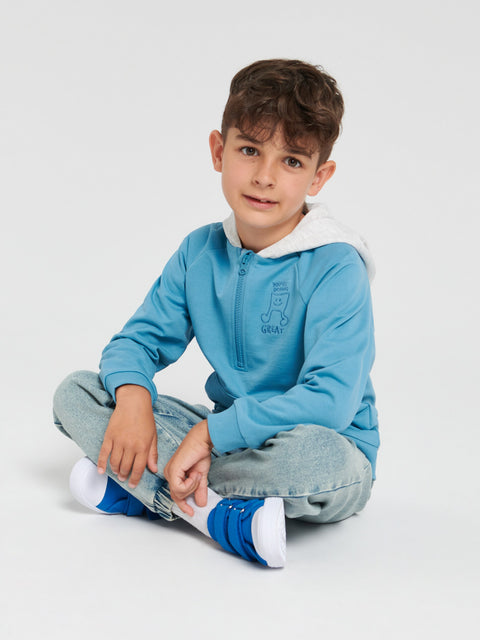 Child wearing a blue sweater with white collar and jeans sitting on a white background