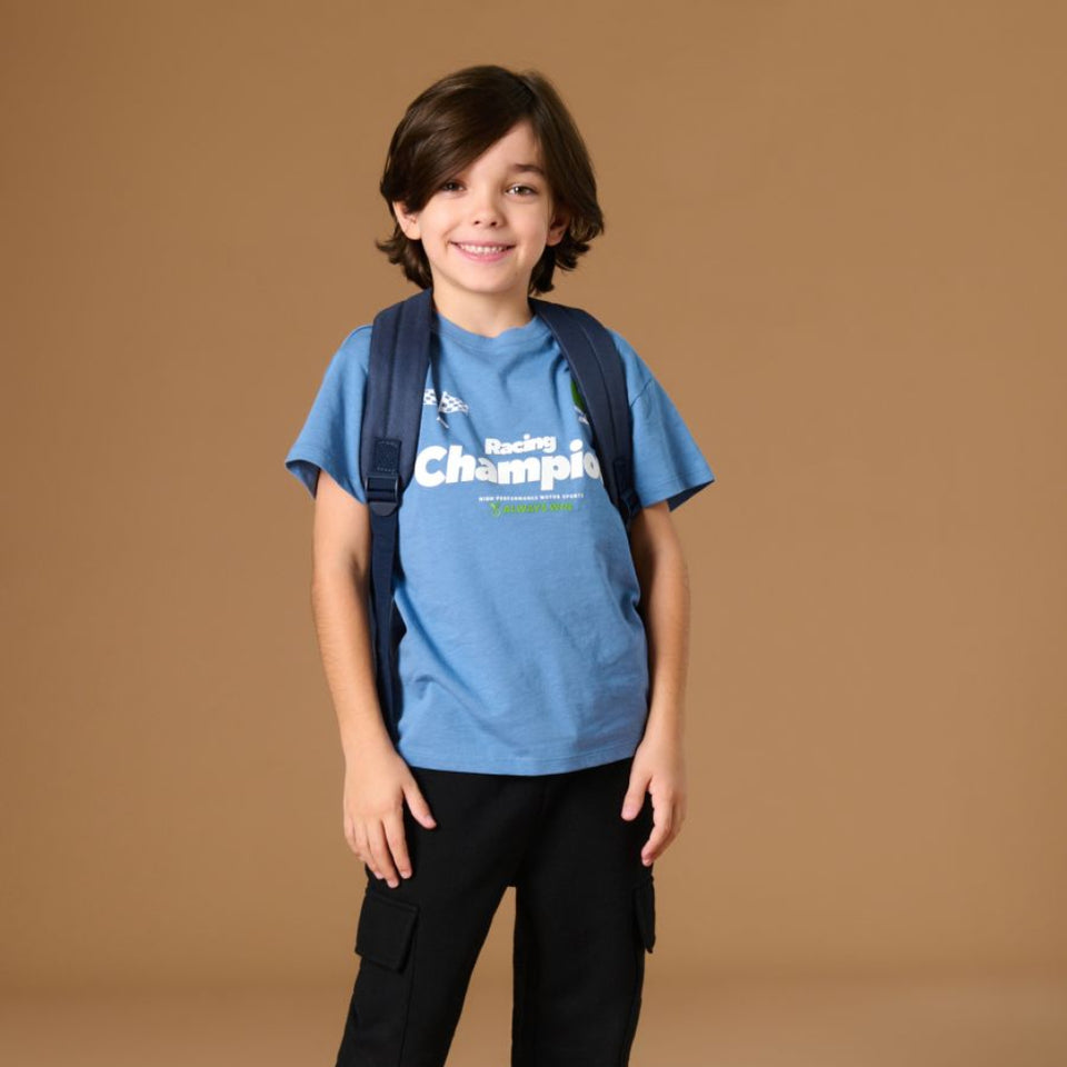 A young boy modeling a blue t-shirt with 'Racing Champions' printed on the front, paired with black cargo pants, black sneakers, and a dark blue backpack, set against a solid tan studio background.