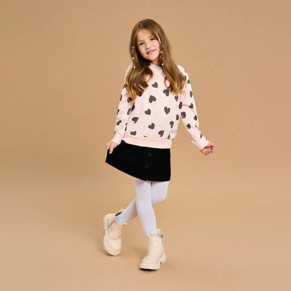 A young girl modeling a light pink sweatshirt with black heart prints, a black mini skirt, white tights, and cream-colored boots, set against a solid beige studio background.