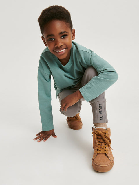 Child wearing a teal long-sleeve shirt and brown shoes on a white background