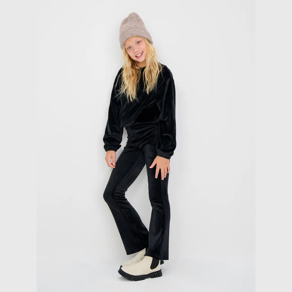 A young girl modeling a black velvet matching lounge set consisting of a long-sleeve top and flared pants, styled with a beige knit beanie and cream slip-on boots against a plain white studio background.