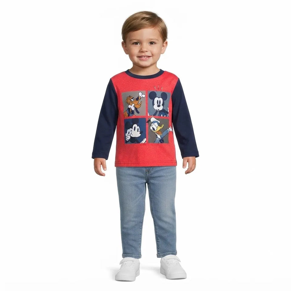 A young child standing against a white background, wearing a red and navy long-sleeve shirt with a Mickey Mouse graphic, blue jeans, and white sneakers.