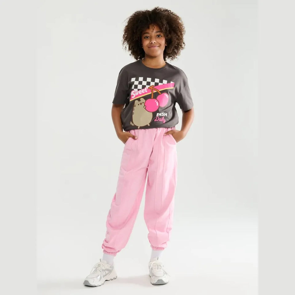A child model wearing a dark gray graphic t-shirt with a Pusheen the cat and pink cherries, paired with light pink jogger pants and white sneakers, standing against a light gray studio background.