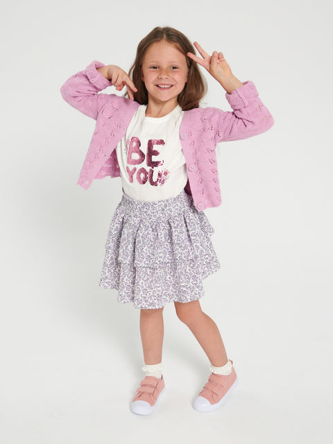 Young girl wearing a pink jacket, white shirt with 'BE YOU' text, and floral skirt on a white background