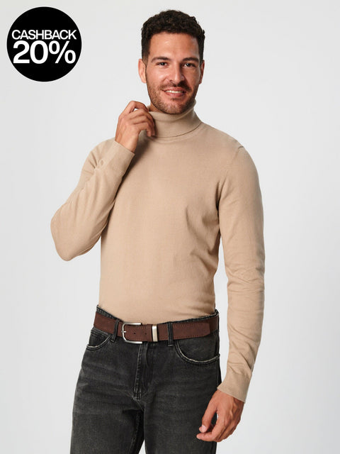 Man wearing a beige turtleneck sweater and dark jeans on a light gray background