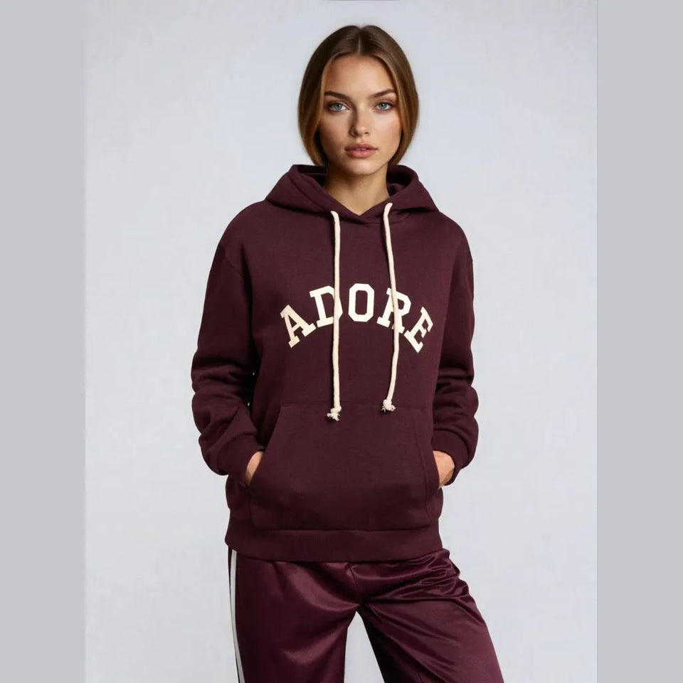 A woman modeling a maroon pullover hoodie with cream drawstrings and a cream 'ADOPE' graphic across the chest, paired with matching maroon satin track pants, against a light gray studio background.