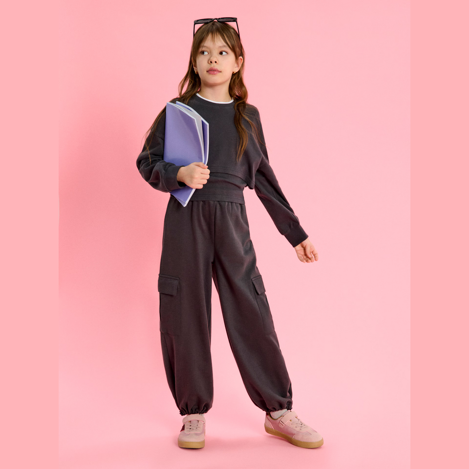 A young girl modeling a dark gray two-piece lounge set consisting of a long-sleeve crop top and matching cargo pants, holding a purple notebook against a solid pink background.