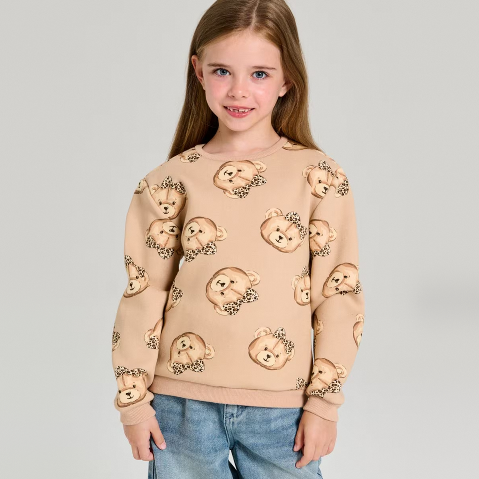 A young girl with long light brown hair wearing a beige crewneck sweatshirt with an all-over teddy bear face pattern and blue jeans, standing against a solid light gray background.