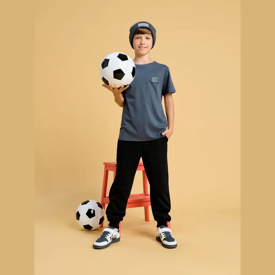 A child model wearing a slate blue t-shirt, black sweatpants, and a gray beanie, holding a soccer ball while standing in front of a beige background with another soccer ball on the floor.