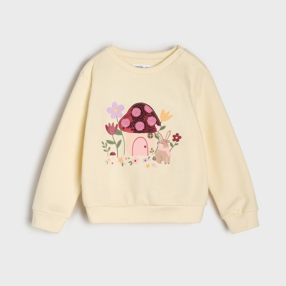 A cream-colored long-sleeve sweatshirt featuring a colorful graphic of a mushroom house, flowers, and a rabbit on the front, displayed against a light gray background.