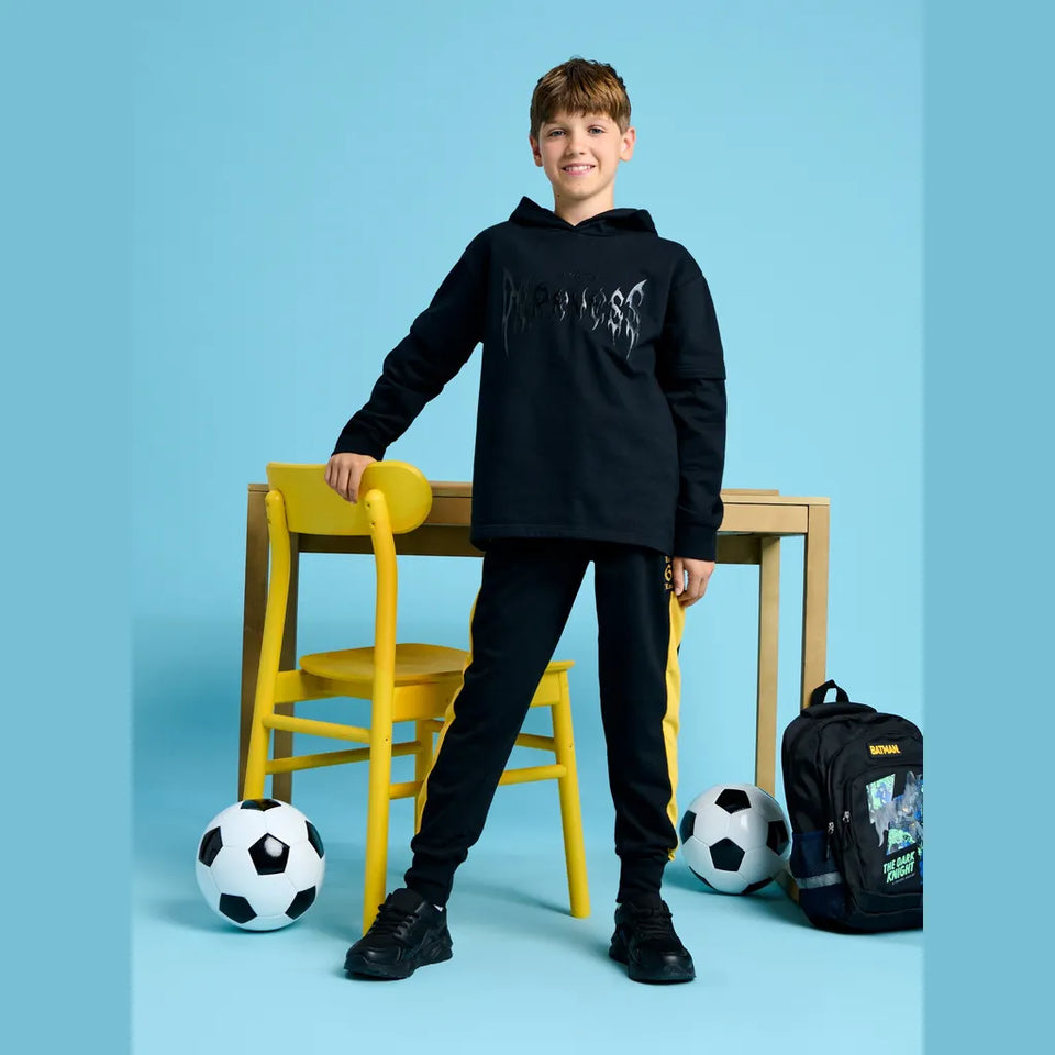 A boy stands in a studio setting wearing a black hoodie and black pants, leaning against a yellow chair beside a wooden table. Soccer balls and a black backpack are on the floor around him against a light blue background.