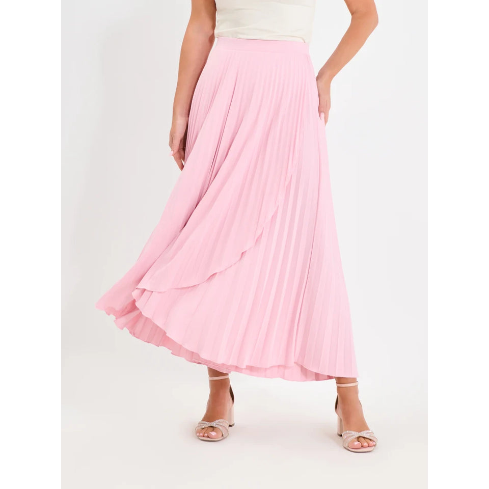 A model wearing a light pink pleated maxi skirt with a layered ruffle hem, paired with a cream top and clear strap sandals, set against a plain white studio background.