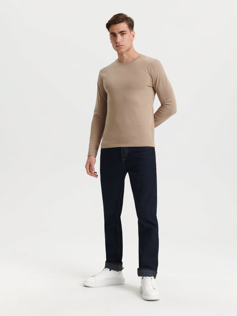 Man wearing a beige sweater and dark jeans on a white background