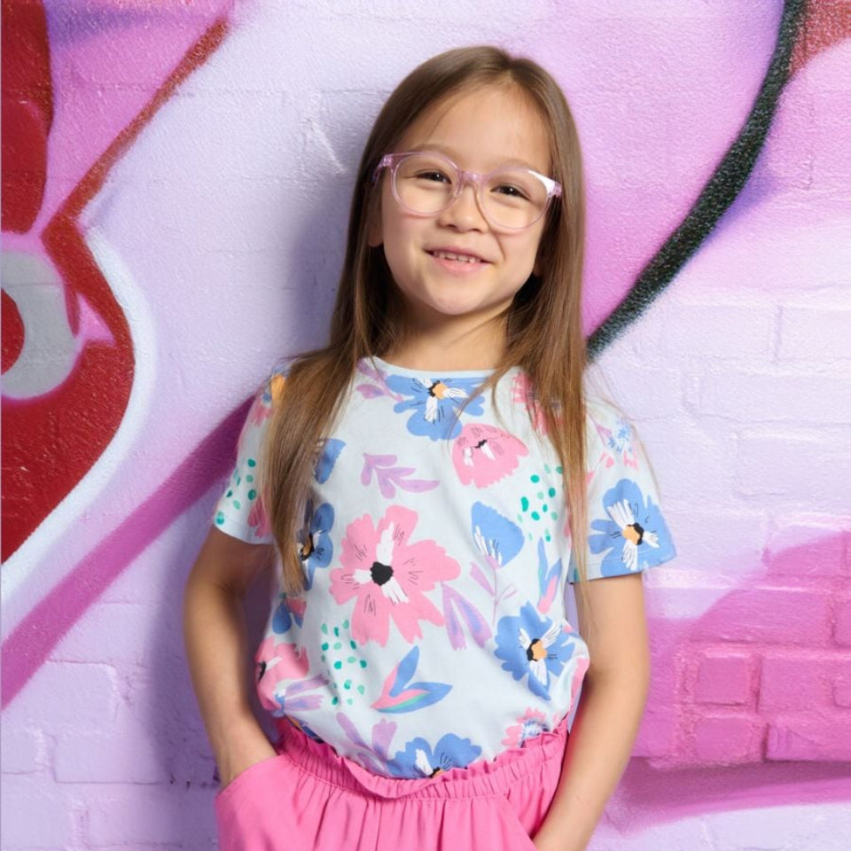 A young girl with long light brown hair and pink-framed glasses stands against a pink and purple brick wall with graffiti, wearing a light blue short-sleeve t-shirt with a pink and blue floral pattern and bright pink shorts.