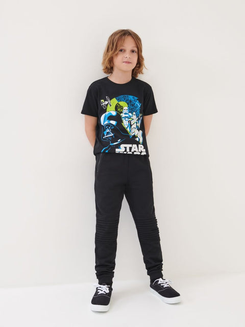 A boy wearing a black short-sleeve T-shirt with a 'Star Wars' character graphic and paired with black trousers and white sneakers.