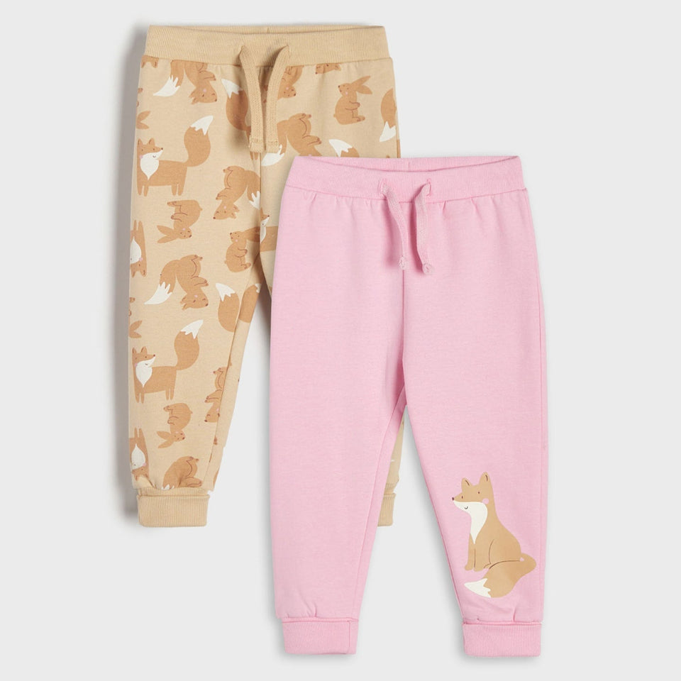 Two pairs of children's jogger sweatpants laid flat on a light gray background. The front pair is light pink with a fox graphic near the lower leg, and the back pair is beige with an allover fox print.