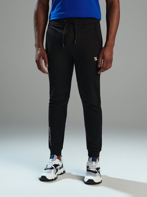 Person wearing black jogger pants with a brand logo, white sneakers, and a blue top on a gray background