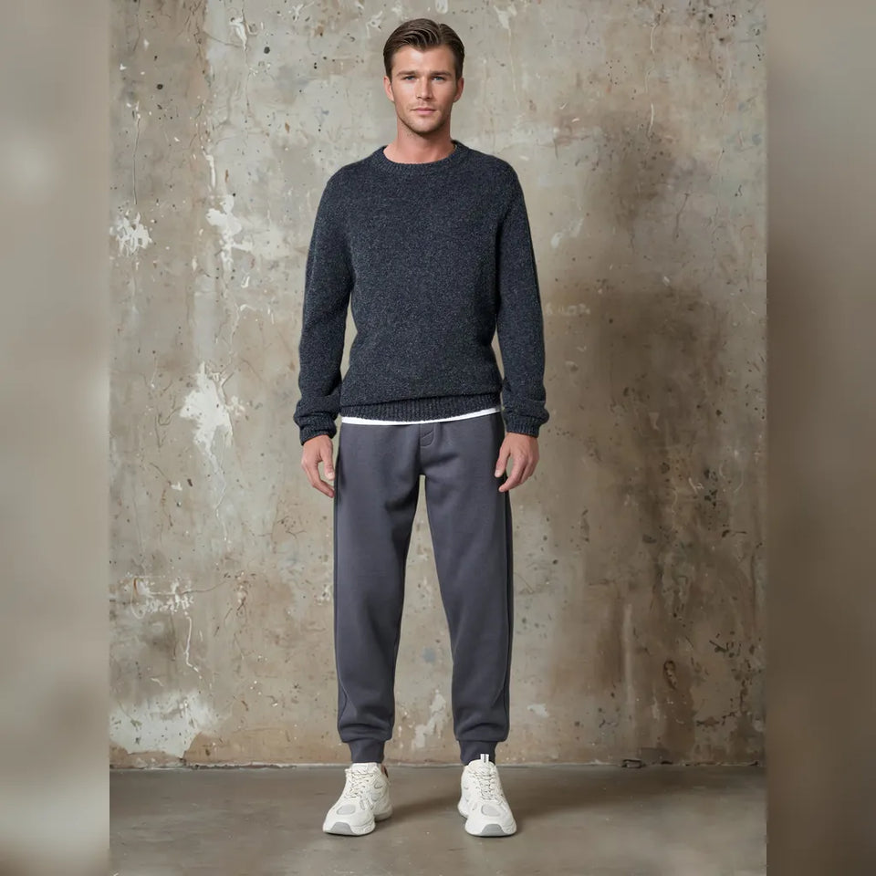 A man modeling a dark heather gray knit crewneck sweater and dark gray sweatpants with white sneakers, standing against a textured concrete wall.