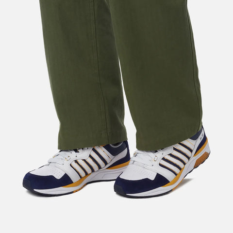 White sneakers with blue and yellow accents worn with green pants on a white background