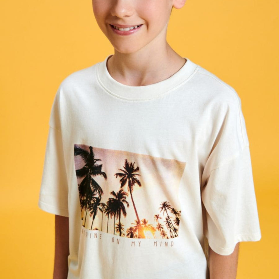A smiling young boy with brown hair wearing a cream-colored t-shirt with a tropical palm tree graphic, set against a solid yellow background.