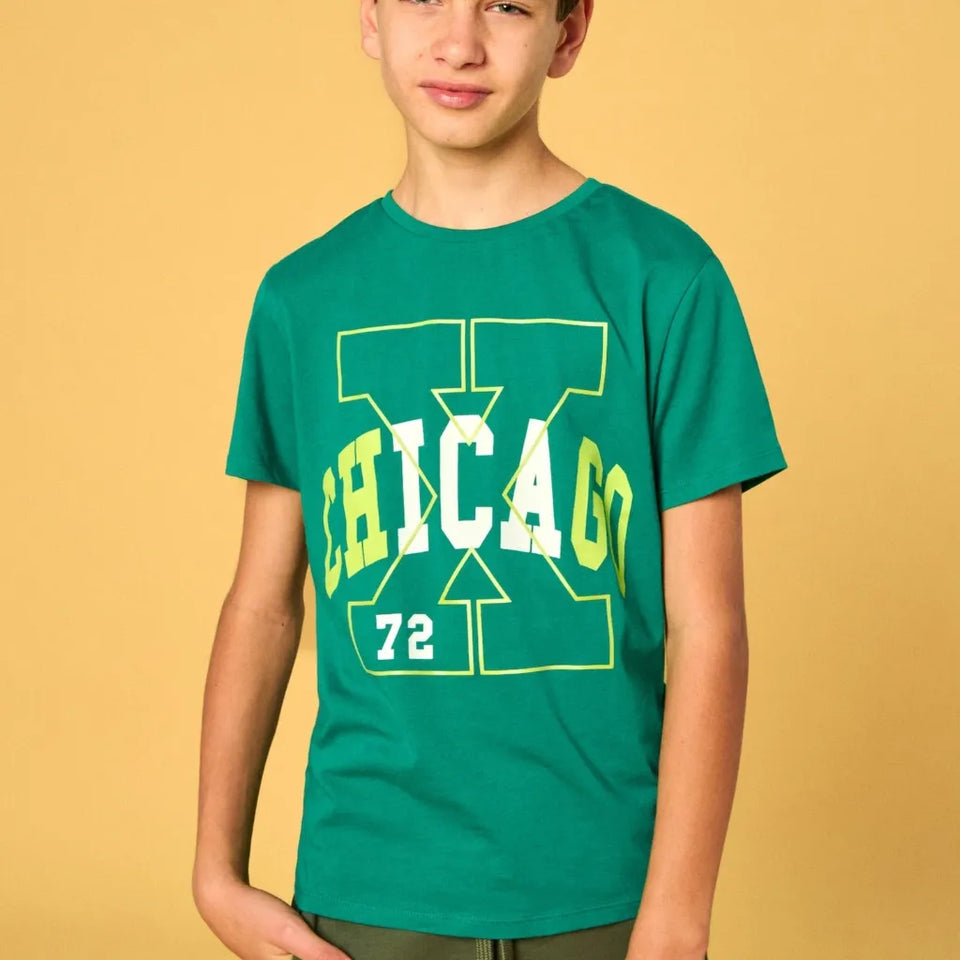 A boy wearing a green short-sleeve t-shirt with a yellow and white graphic on the front, standing against a solid yellow background.