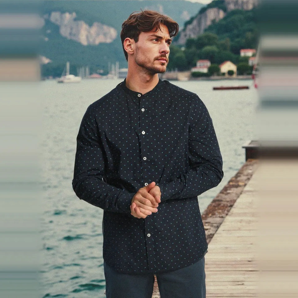 A man wearing a dark navy long-sleeve button-up shirt with a small white geometric pattern, standing on a wooden dock with a scenic lake and mountains in the background.