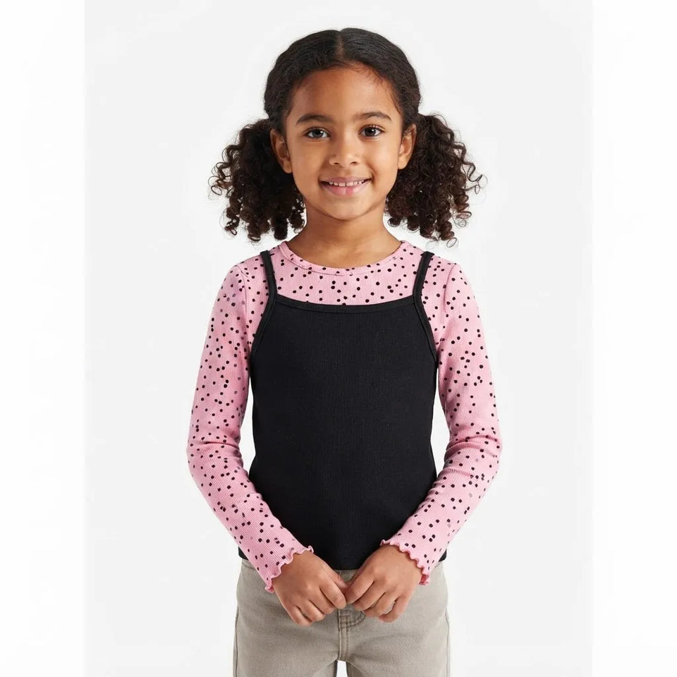 A young girl with curly dark hair is wearing a two-piece long-sleeve shirt set. The top features a black front panel with shoulder straps, layered over a pink long-sleeve shirt with small black polka dots.