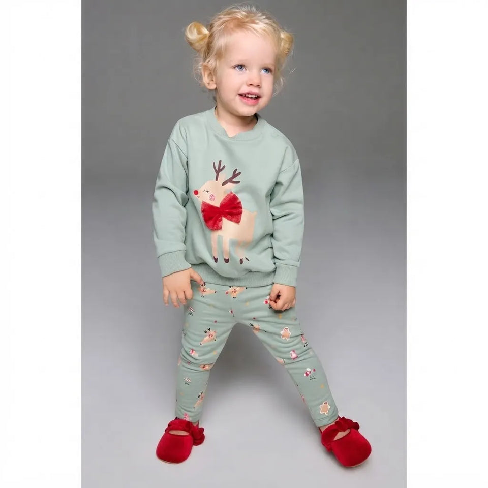 A toddler girl wearing a sage green two-piece pajama set with a red bow-tied reindeer on the sweatshirt and small floral prints on the pants, paired with red slippers, standing against a gray studio background.