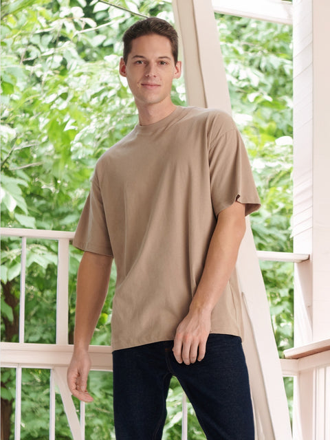 Man wearing a beige t-shirt and dark pants standing on a balcony with greenery in the background