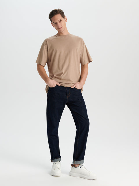 Man wearing a beige t-shirt and dark jeans on a white background