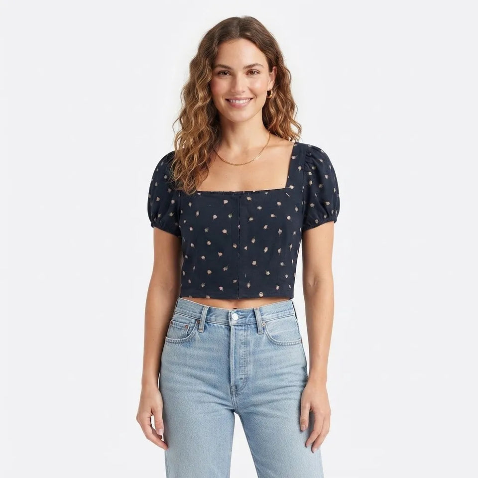 A woman modeling a navy blue puff-sleeve crop top with a beige and white polka dot pattern, paired with light-wash blue jeans, against a plain light gray background.