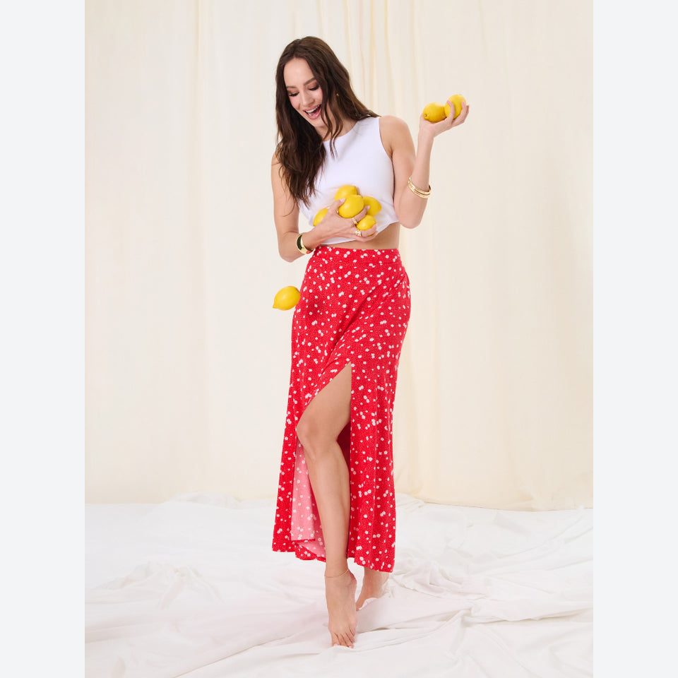 A woman stands barefoot on a white bed against a cream curtain backdrop, wearing a red wrap-style maxi skirt with a white floral pattern and a high side slit. She holds several yellow lemons in both hands.