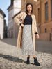 Woman in a beige coat and patterned skirt standing on a cobblestone street.