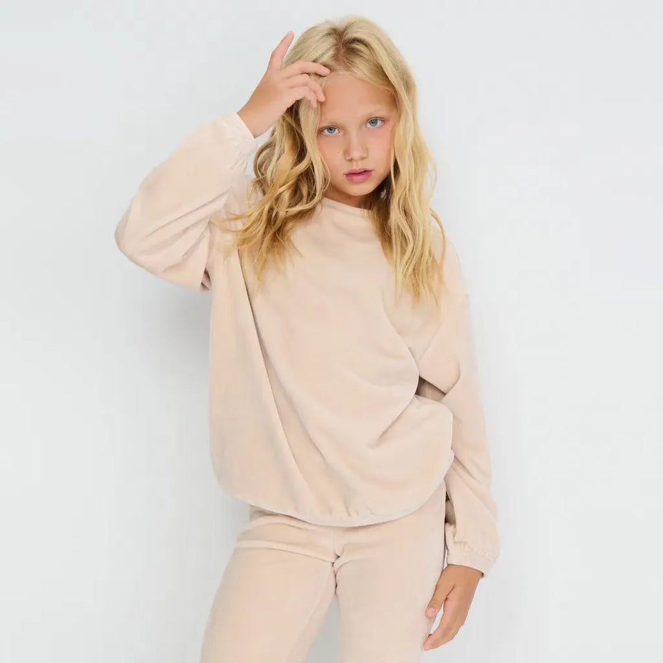 A young girl with blonde hair wearing a matching beige sweatshirt and sweatpants set, posing against a plain white studio background.