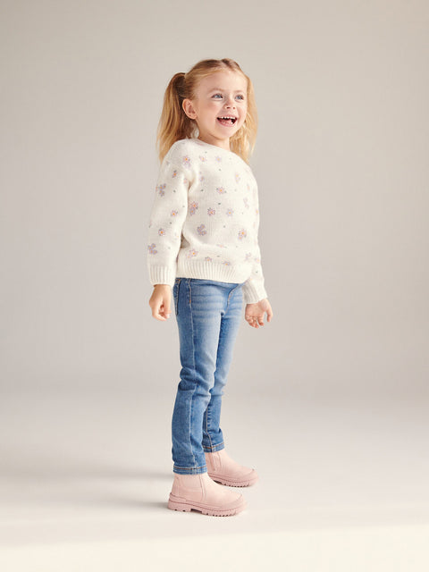 Young girl wearing a white sweater with floral patterns, blue jeans, and pink boots on a light gray background
