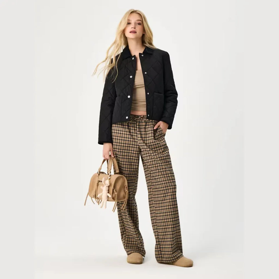 A blonde model stands against a light gray studio background wearing a black quilted jacket over a beige ribbed top, wide-leg brown and beige plaid trousers, and tan shoes, while holding a small tan handbag.