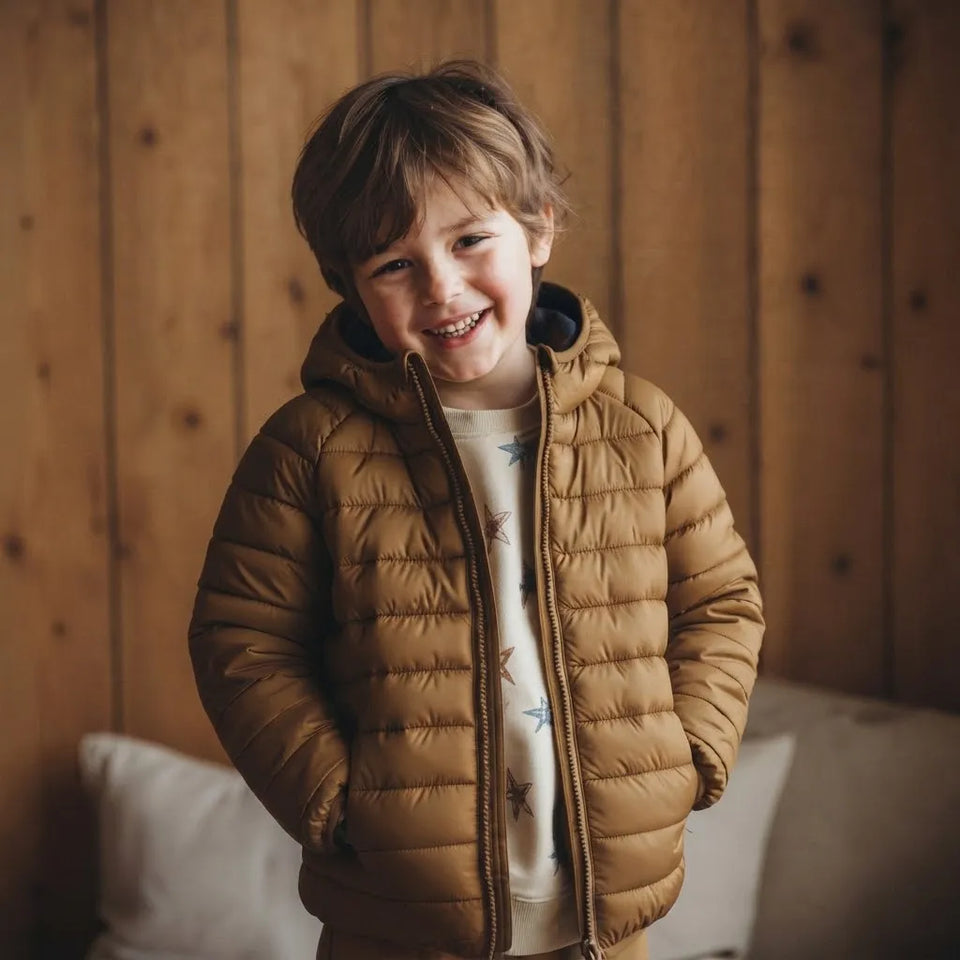 A young boy smiling while wearing a tan quilted puffer jacket with a hood over a white star-patterned shirt, standing against a warm wood-paneled wall.