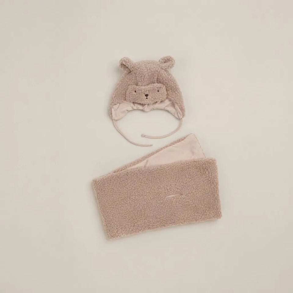 A soft fleece baby accessory set in a dusty blush pink color, featuring a bear-face aviator hat with ear flaps and a matching rectangular headband, laid out on a light beige background.