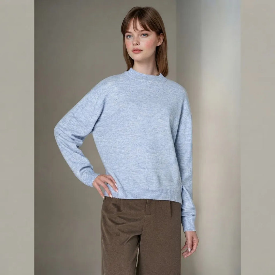 A woman modeling a light blue heathered knit sweater with a mock neck and long sleeves, paired with brown trousers, against a neutral beige studio background.
