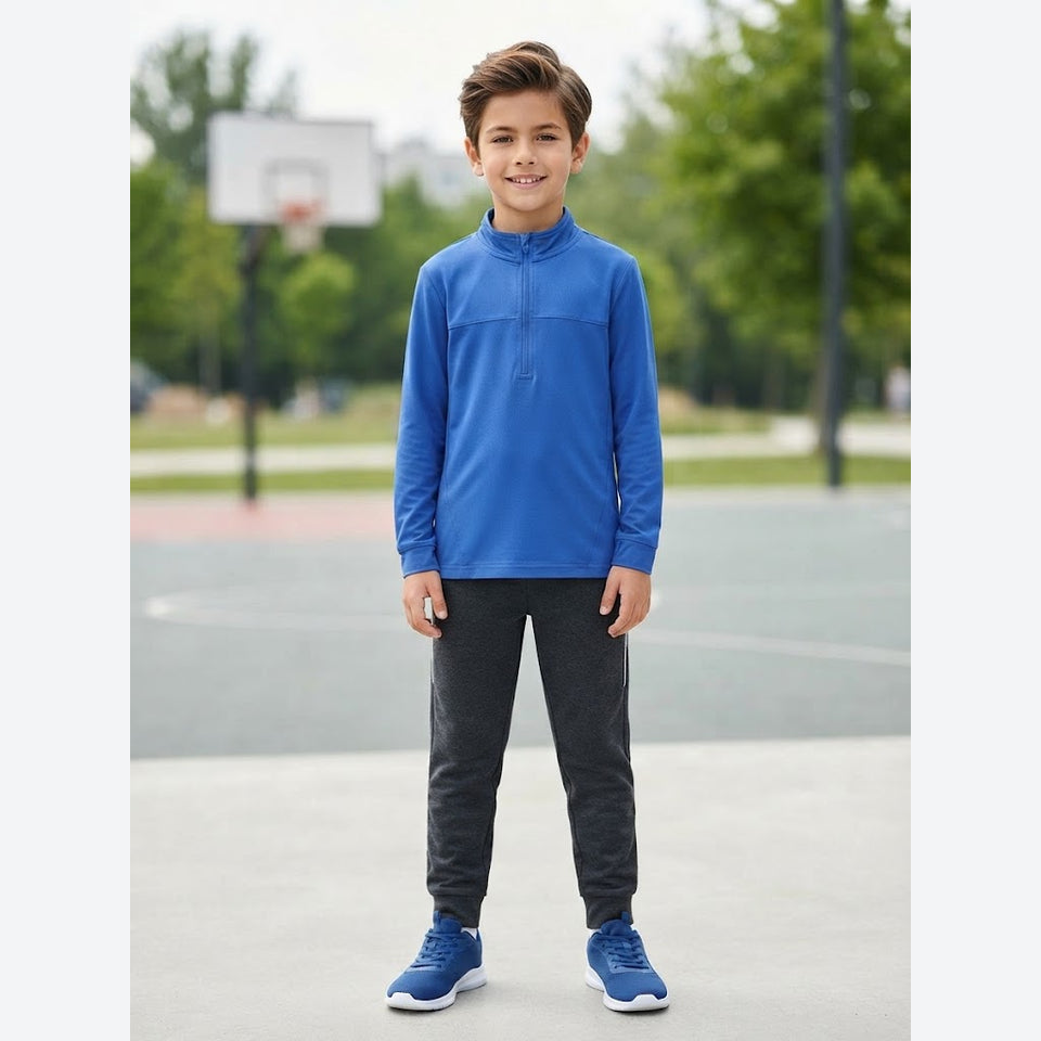 A young boy stands on an outdoor basketball court wearing a bright blue long-sleeve quarter-zip pullover, dark gray jogger pants, and blue athletic sneakers.