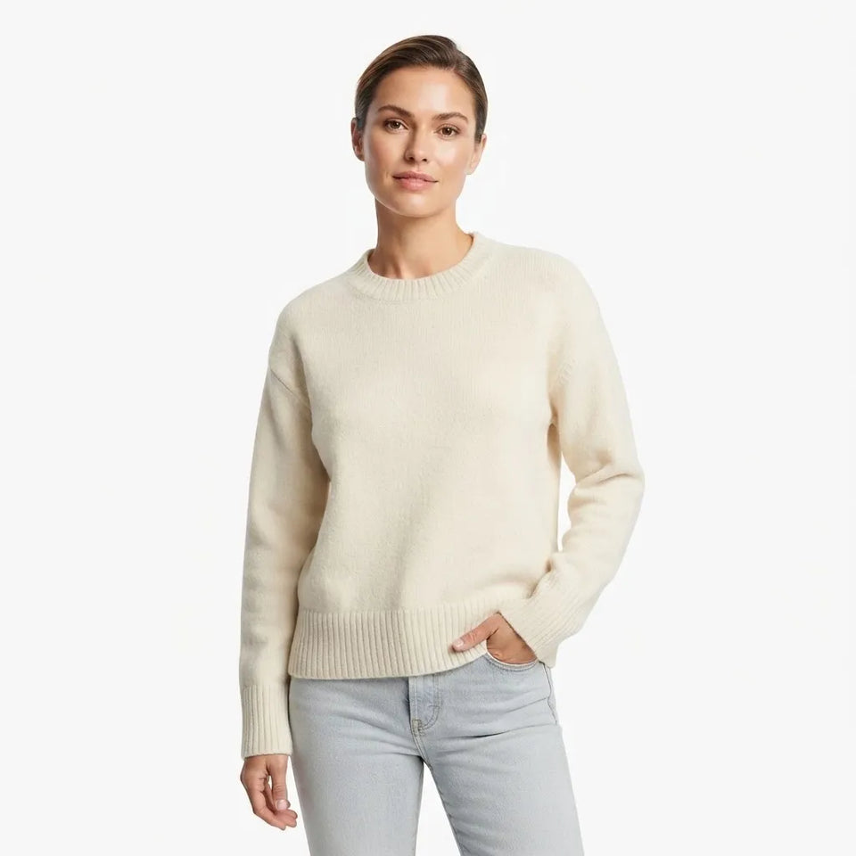 A woman modeling a cream-colored crewneck knit sweater with light-wash jeans against a plain white studio background.