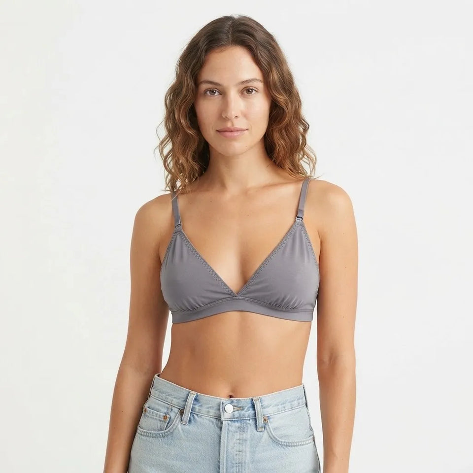 A woman wearing a gray triangle bralette with thin straps and a twisted front detail, paired with light-wash blue jeans, against a plain white studio background.