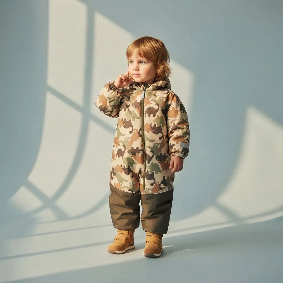 A toddler stands in a studio wearing a hooded camouflage snowsuit with brown knee panels and tan boots, set against a pale blue wall with window shadow patterns.
