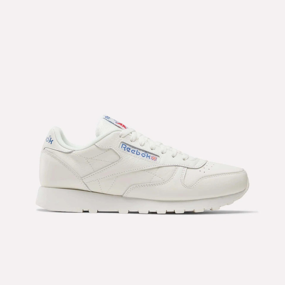 A classic Reebok sneaker in off-white leather, shown from the side against a light gray background. The shoe features white laces, a thick white midsole, and blue and red accents on the Reebok branding on the side and heel.