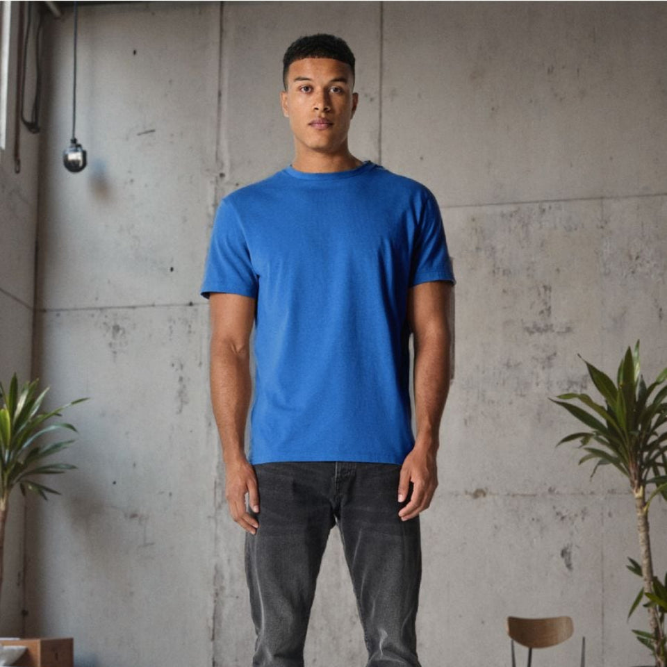 A man wearing a bright blue crewneck t-shirt, dark gray jeans, and black sneakers, standing in a modern industrial room with a concrete wall and plants.