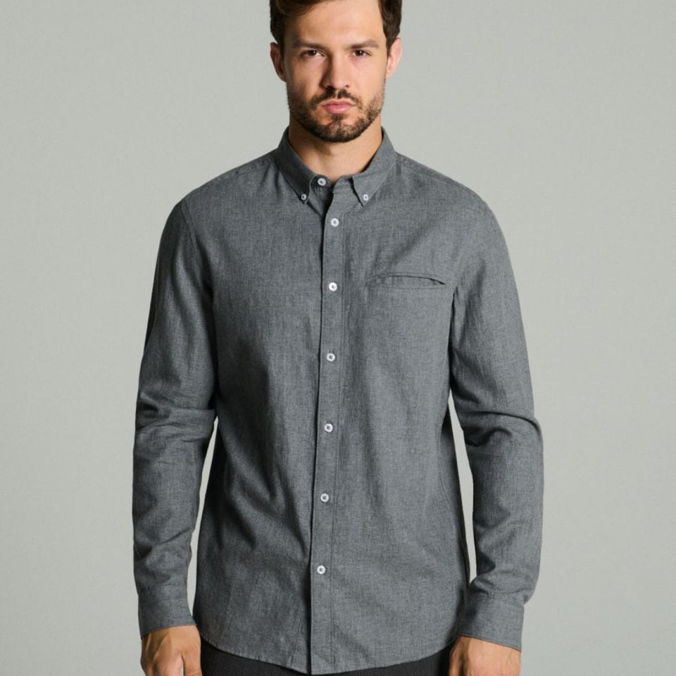 A man wearing a dark gray long-sleeve button-down shirt with a chest pocket, paired with dark trousers, standing against a solid light gray background.