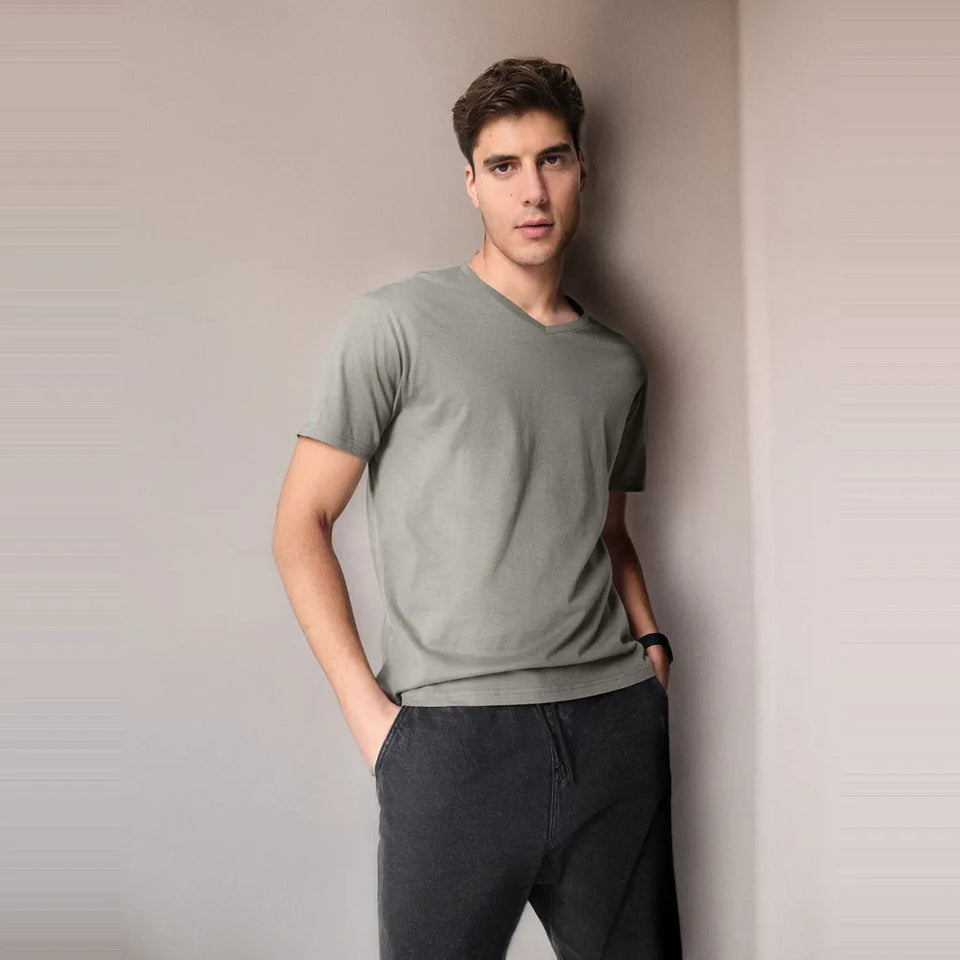 A man wearing a light sage green v-neck t-shirt and dark gray pants, standing against a neutral light gray wall.